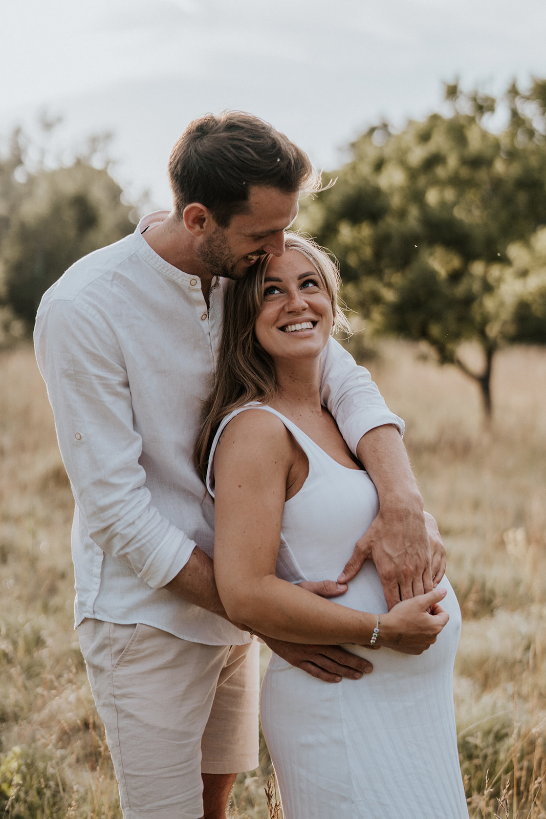 Werdende Eltern in der Abendsonne, der Mann umarmt den Babybauch seiner Partnerin