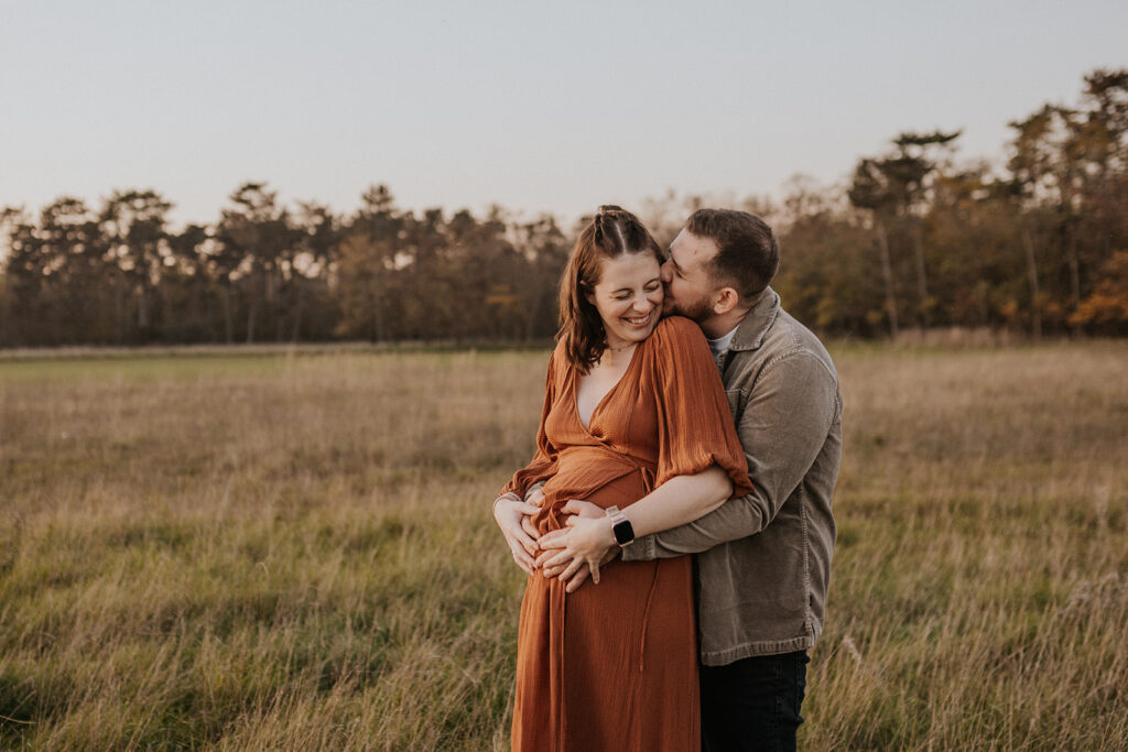 Werdende Eltern stehen im Feld, sie im rostroten Kleid mit Haenden am Babybauch