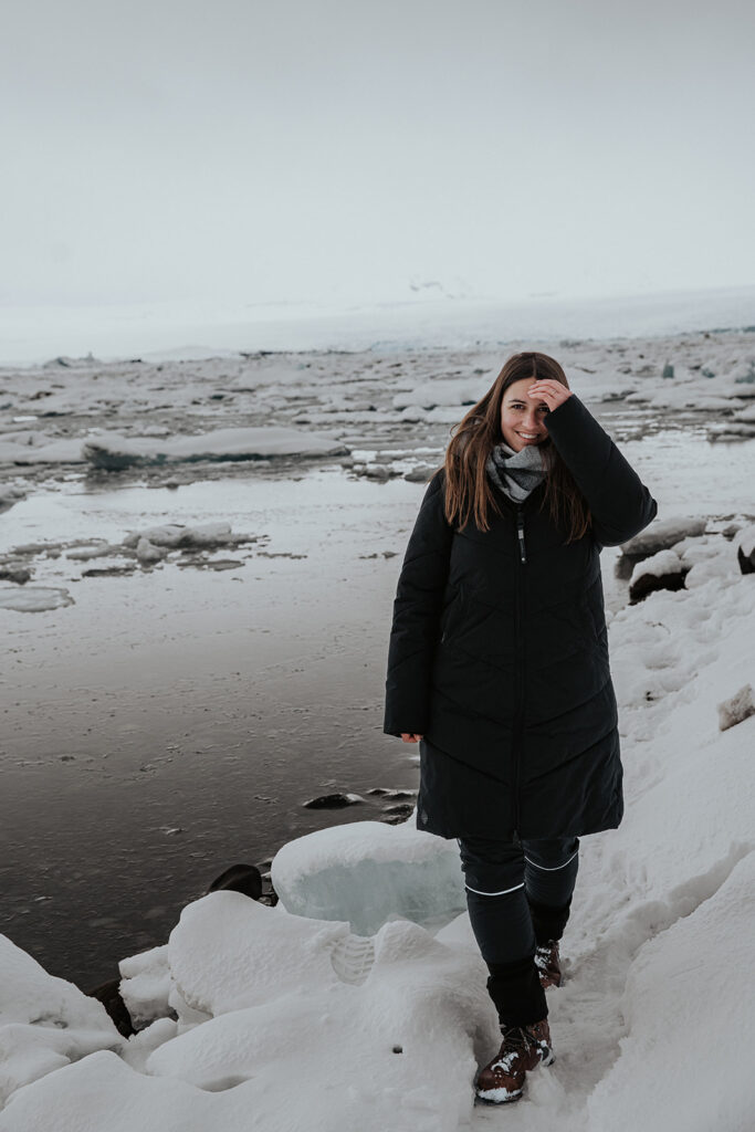 Frau im Wintermantel steht am Ufer einer Gletscherlagune in Island