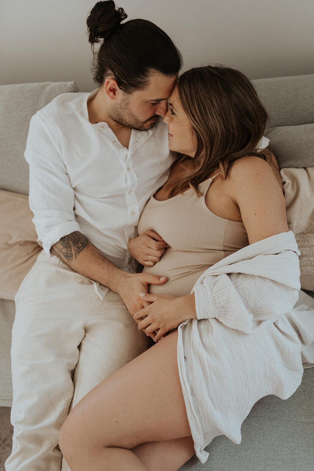Werdende Eltern liegen Nase an Nase auf dem Sofa, die Hände gemeinsam auf dem Babybauch