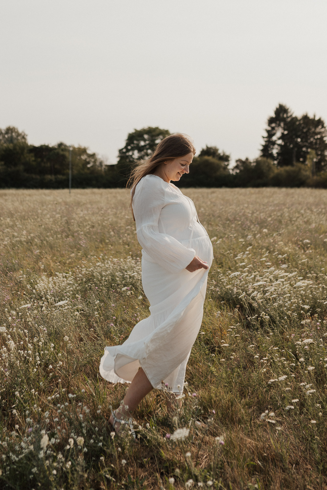 Schwangere im weißen Kleid geht durch eine sommerliche Wiese